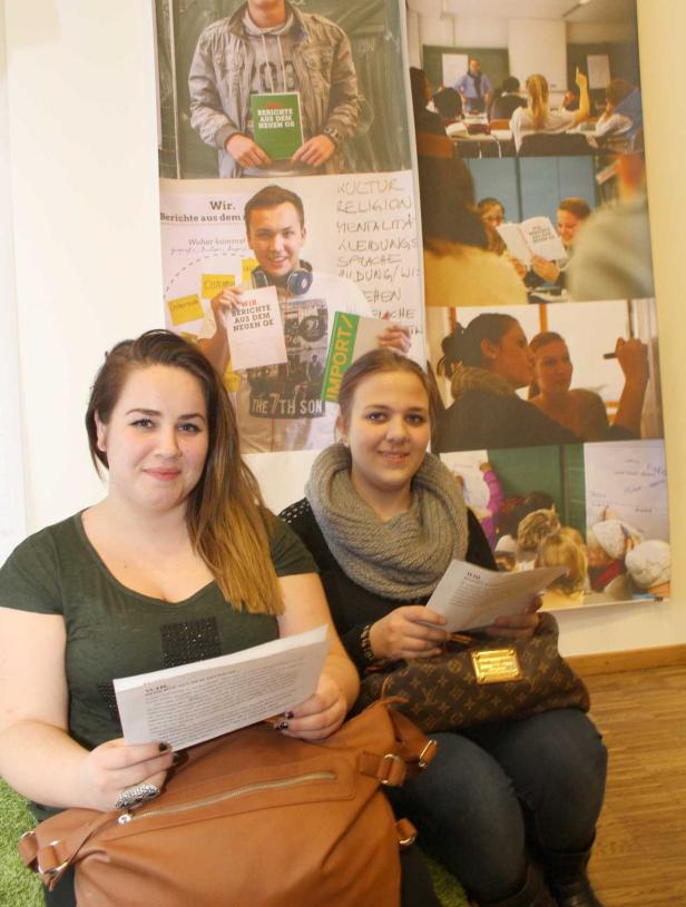 Zwei junge Frauen sitzen vor einem Poster mit dem Titel „Wir. Berichte aus dem neuen OE“.