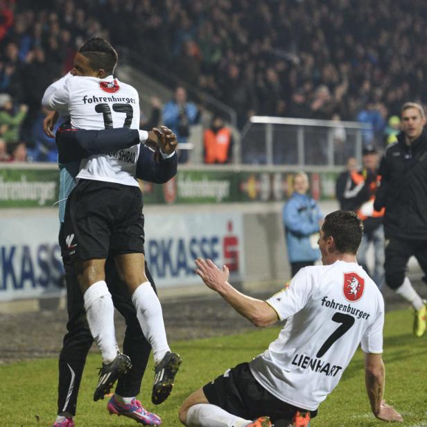 Zwei Fußballspieler feiern ein Tor, einer liegt auf dem Rasen, der andere springt in die Arme eines Trainers.