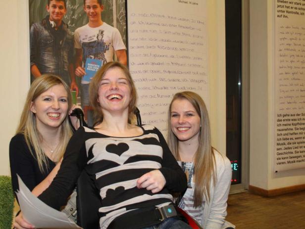 Drei junge Frauen posieren lachend für ein Foto, eine davon im Rollstuhl.