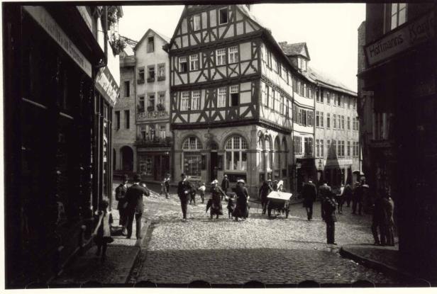 Eine belebte Straße mit Kopfsteinpflaster und Fachwerkhäusern in einer deutschen Stadt um 1900.