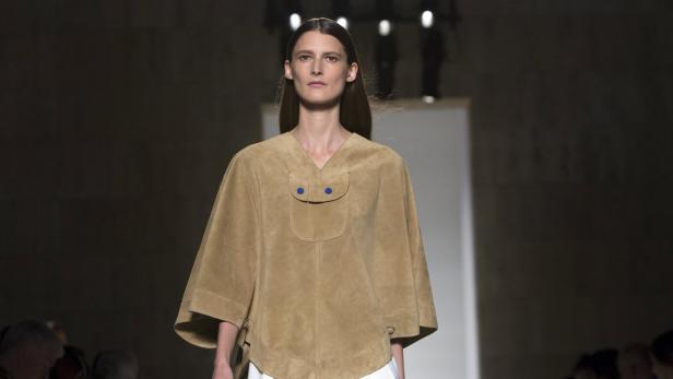Eine Frau präsentiert ein beiges Wildleder-Cape mit blauen Knöpfen auf einem Laufsteg.