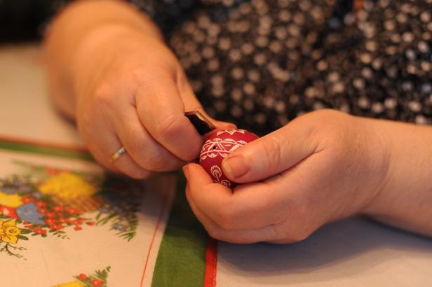 Eine Person verziert ein rotes Osterei mit einem Schnitzwerkzeug.