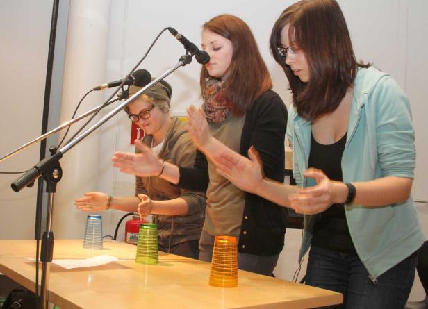 Drei junge Frauen stehen an einem Tisch und machen Cup-Percussion mit Plastikbechern.
