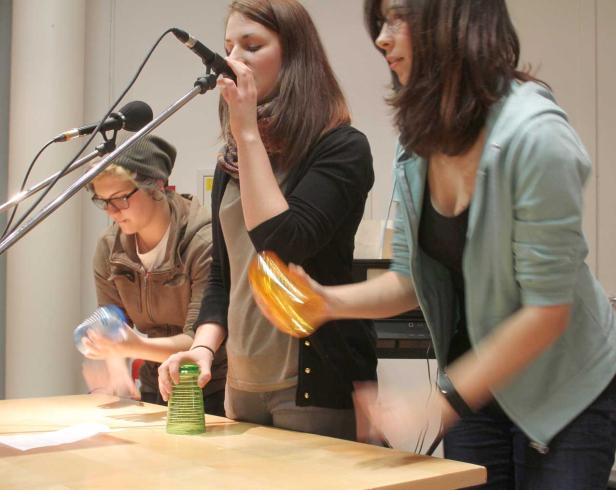 Drei Frauen performen mit Bechern und Mikrofonen auf einer Bühne.
