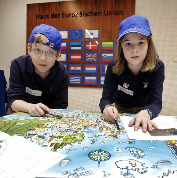 Zwei Kinder mit blauen Kappen betrachten eine Europakarte vor dem „Haus der Europäischen Union“.