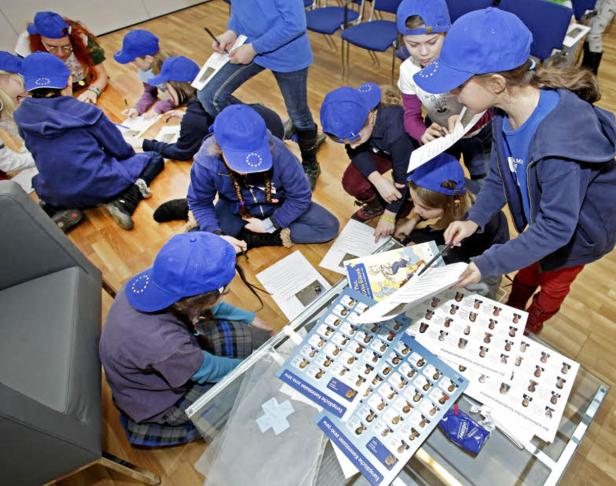 Eine Gruppe Kinder mit blauen Kappen sitzt auf dem Boden und arbeitet an Aufgaben zum Thema Europäische Kommission.