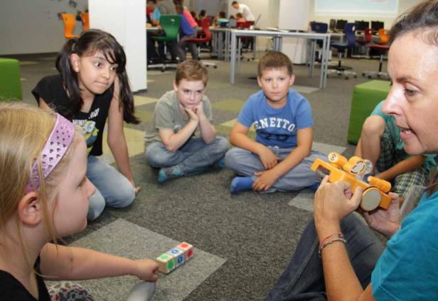 Eine Gruppe Kinder sitzt im Kreis und lernt mit einer Lehrerin spielerisch Programmieren.