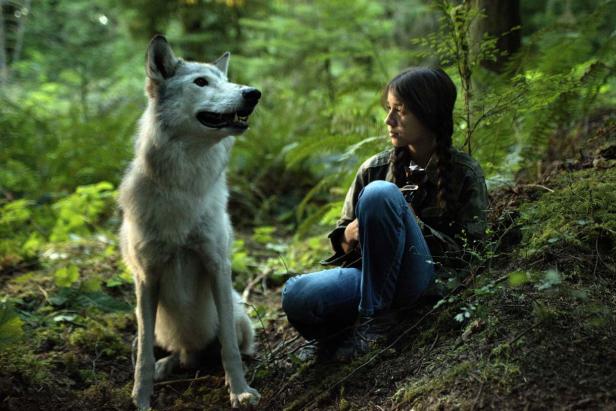 Eine junge Frau sitzt im Wald neben einem Wolf.