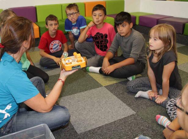 Eine Frau erklärt einer Gruppe von Kindern einen kleinen gelben Roboter.