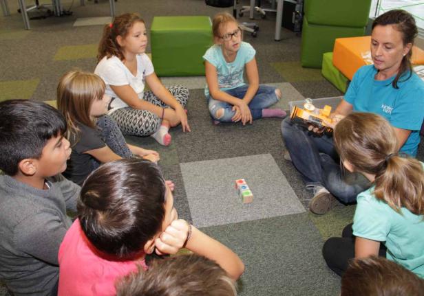 Eine Gruppe Kinder sitzt mit einer Betreuerin im Kreis und betrachtet einen Roboter.
