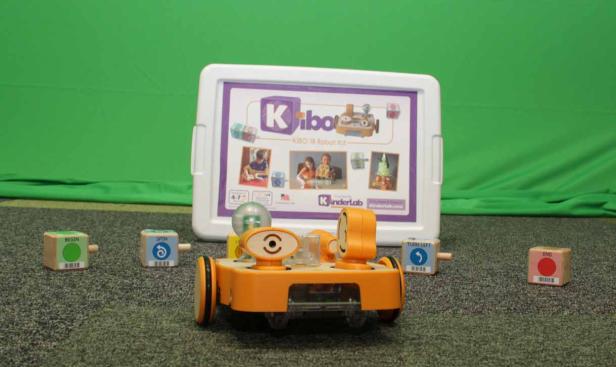 Ein orangefarbener KIBO-Roboter steht vor einer Kiste und mehreren Programmierblöcken.