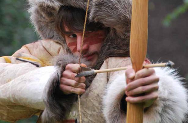 Ein Mann mit Gesichtsbemalung zielt mit Pfeil und Bogen.