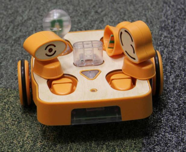 Ein orangefarbener Roboter mit Rädern und Sensoren auf einem grauen Teppich.