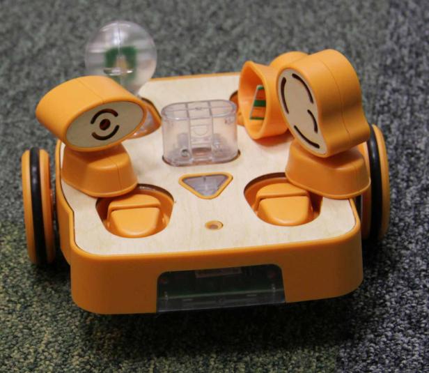 Ein kleiner, orangefarbener Roboter mit Rädern und Sensoren auf einem Teppich.