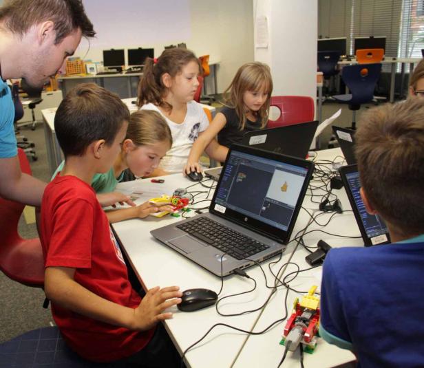 Eine Gruppe Kinder sitzt mit einem Lehrer an Tischen und programmiert Roboter mit Laptops.