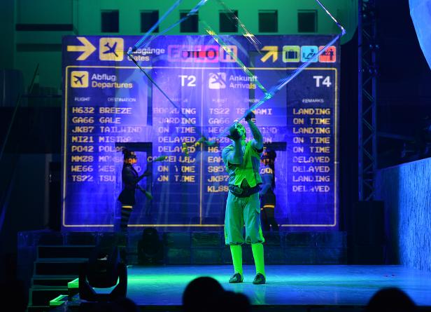 Ein Mann in grüner Kleidung steht vor einer Anzeigetafel mit Flugdaten auf einer Bühne.