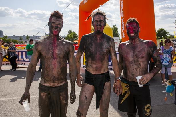 Drei Männer, mit Schlamm bedeckt, stehen nach einem Hindernislauf vor einem orangefarbenen Torbogen.