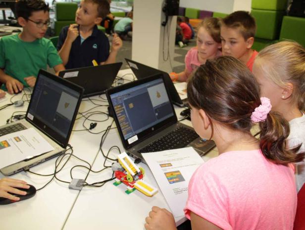 Eine Gruppe Kinder arbeitet mit Laptops und Lego-Robotern an einem Tisch.