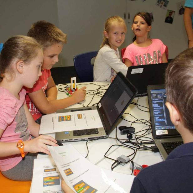 Eine Gruppe Kinder sitzt an einem Tisch und arbeitet mit Laptops und Lego-Robotern.
