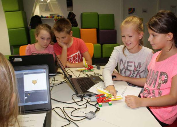 Eine Gruppe Kinder arbeitet mit Laptops und Lego-Robotern an einem Tisch.