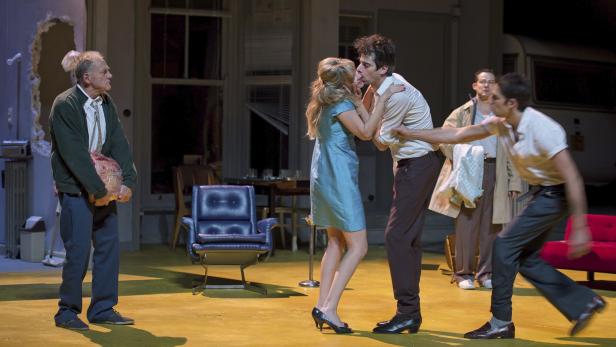 Auf einer Theaterbühne umarmen sich ein Mann und eine Frau, während andere Schauspieler zusehen.