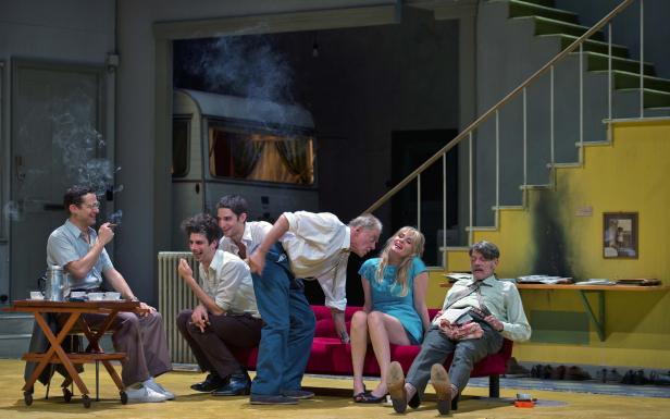 Eine Theaterszene mit sechs Schauspielern in einem Wohnzimmer mit Wohnwagen im Hintergrund.