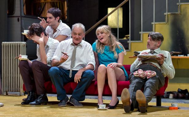 Eine Gruppe von Schauspielern sitzt auf einer roten Couch und raucht Zigarren während einer Theateraufführung.