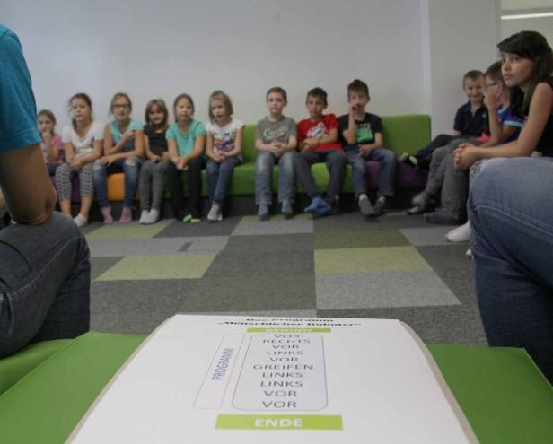 Eine Gruppe Kinder sitzt auf einer Bank, im Vordergrund ein Blatt Papier mit Anweisungen für einen Roboter.