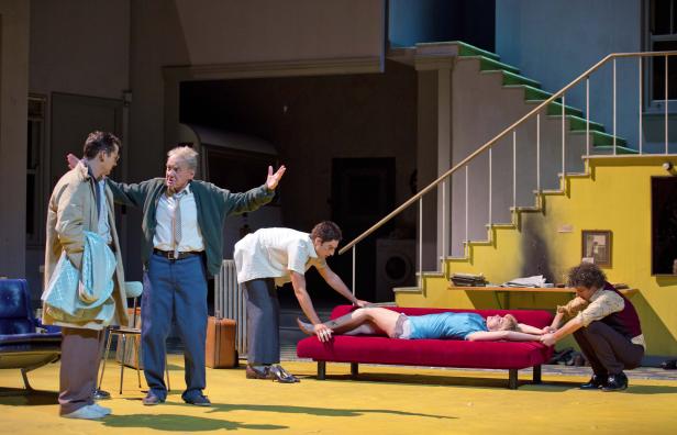 Auf einer Theaterbühne kümmern sich vier Personen um eine bewusstlose Frau auf einer roten Couch.