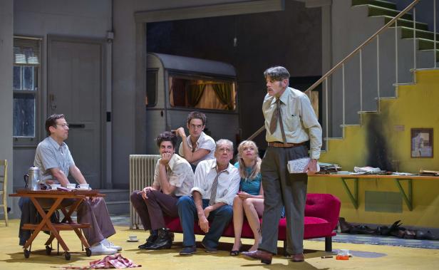 Eine Gruppe von Schauspielern auf einer Theaterbühne vor einem Wohnwagen.