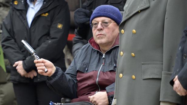 Ein Mann mit Brille und Mütze sitzt im Rollstuhl und hält ein Mikrofon.