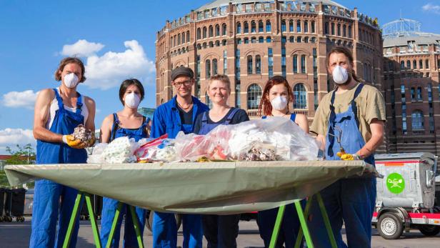 Eine Gruppe von Menschen mit Masken steht hinter einem Tisch voller Müll vor der Kulisse der Wiener Gasometer.