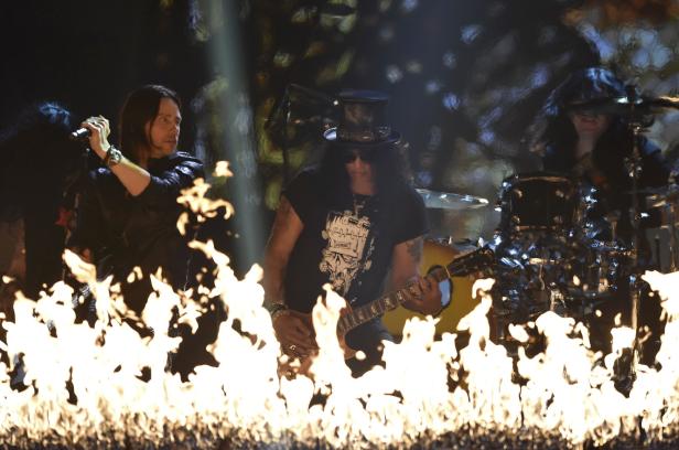 Slash spielt Gitarre vor einer Feuerwand, während ein Sänger und ein Schlagzeuger im Hintergrund zu sehen sind.