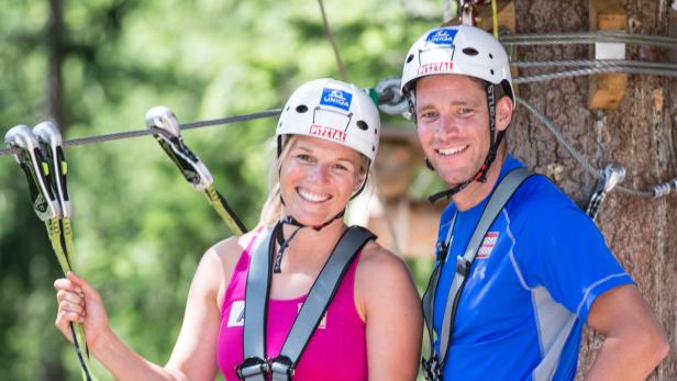 Zwei Personen mit Helmen und Sicherheitsausrüstung lächeln auf einem Hochseilgarten in die Kamera.