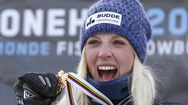 Eine jubelnde blonde Frau mit einer Goldmedaille um den Hals und einer blauen Mütze auf dem Kopf.