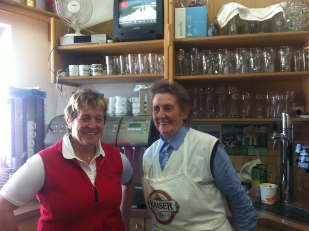 Zwei Frauen stehen lächelnd in einer Gastronomieküche vor Regalen voller Gläser.