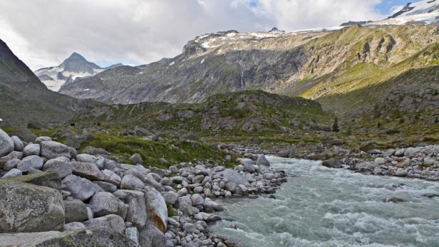 Ein Gebirgsbach fließt durch eine steinige Landschaft mit schneebedeckten Gipfeln im Hintergrund.