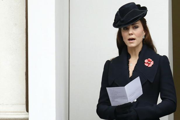 Kate Middleton mit Hut und Mohnblume liest aus einem Dokument.
