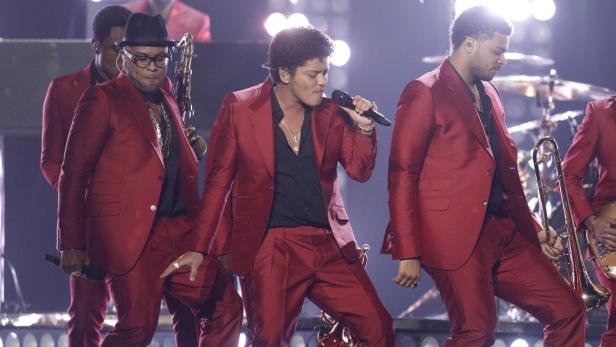Bruno Mars tritt mit seiner Band in roten Anzügen auf einer Bühne auf.