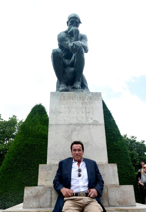 Arnold Schwarzenegger sitzt vor der Statue „Der Denker“ von Rodin.