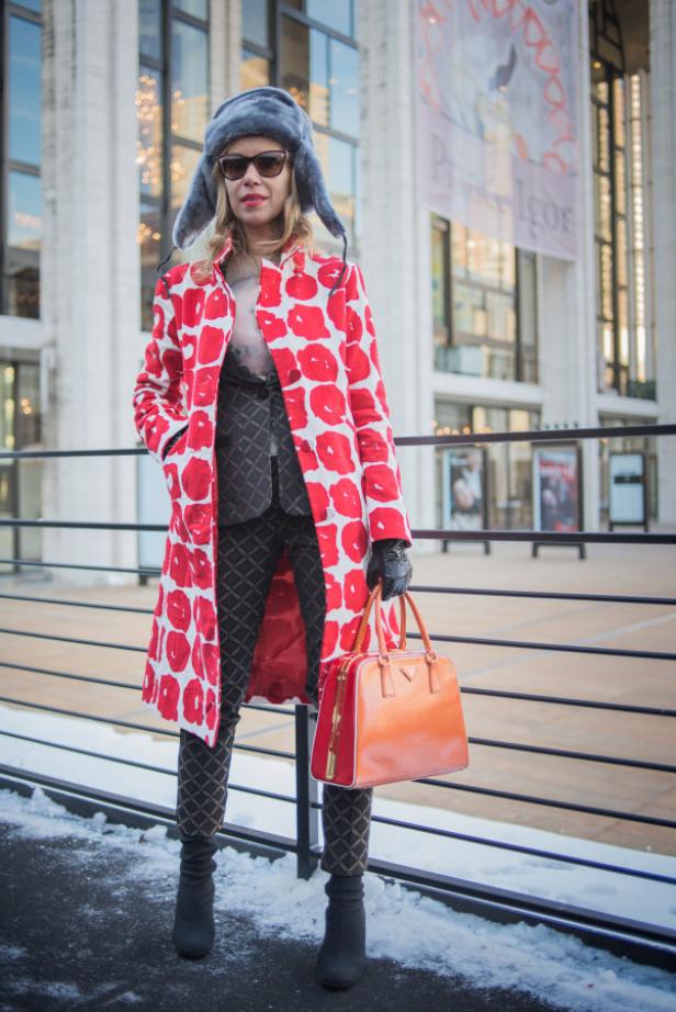 Eine Frau mit Pelzmütze, rotem Mantel und orangefarbener Prada-Tasche steht vor einem Gebäude.