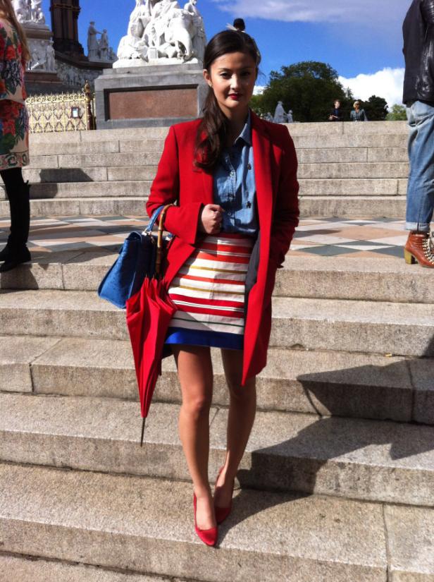 Eine Frau in einem roten Mantel und gestreiften Rock steht auf einer Steintreppe.