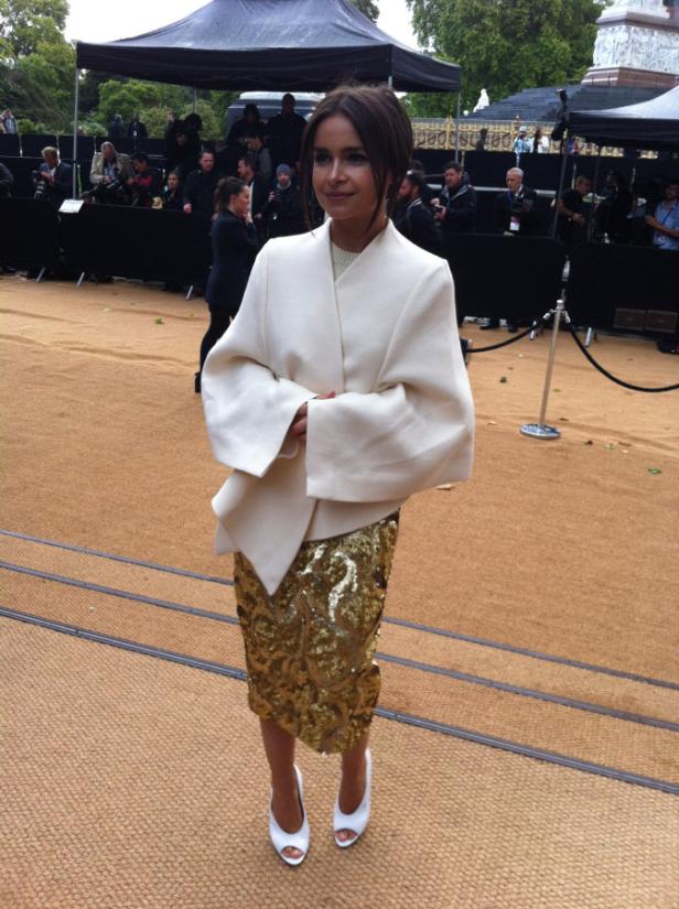 Eine Frau in einem weißen Blazer und einem goldenen Paillettenrock steht auf einem roten Teppich.