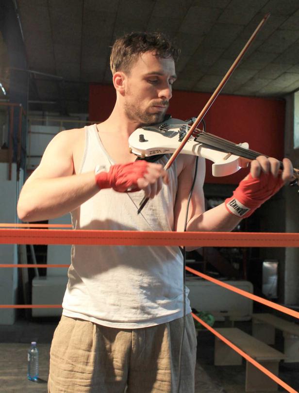 Ein Mann mit Bandagen an den Händen spielt Geige in einem Boxring.