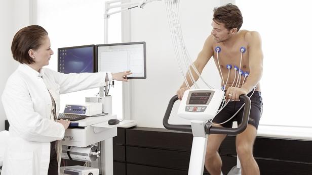 Ein Mann unterzieht sich einem Belastungs-EKG auf einem Fahrradergometer, überwacht von einer Ärztin.