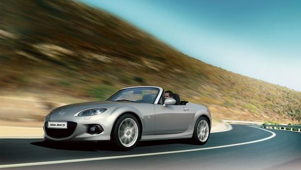 Ein silberner Mazda MX-5 fährt auf einer kurvenreichen Straße entlang eines Hügels.
