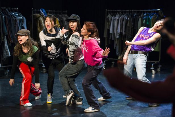 Eine Gruppe junger Leute tanzt auf einer Bühne vor einem Kleiderständer.