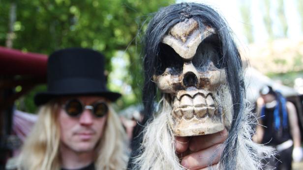 Eine Person hält eine Totenkopfmaske mit Haaren vor dem Gesicht, im Hintergrund eine weitere Person mit Zylinder.