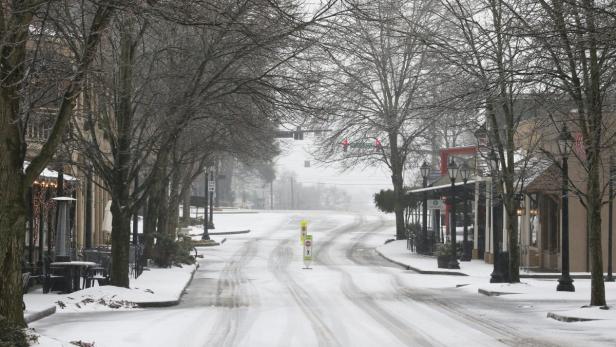 Eine verschneite Straße in einer Kleinstadt mit Bäumen und Geschäften an den Seiten.