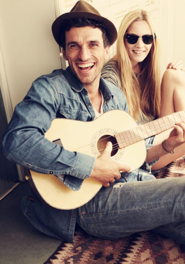 Ein lächelnder Mann mit Hut spielt Gitarre, während eine Frau mit Sonnenbrille daneben sitzt.